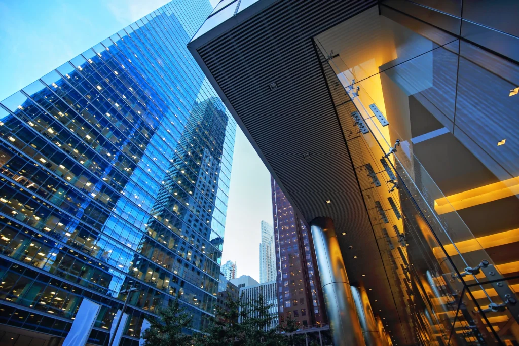 This image captures the dynamic architecture of a modern financial district at dusk. Tall glass buildings tower above, reflecting the blue hues of the sky and the lights from surrounding structures. The perspective is upward, showing the juxtaposition of glass and steel that characterizes urban development. The design features sleek, reflective surfaces and sharp angles, creating a sense of grandeur and corporate elegance. The image conveys the bustling activity and architectural beauty of a city's financial hub.